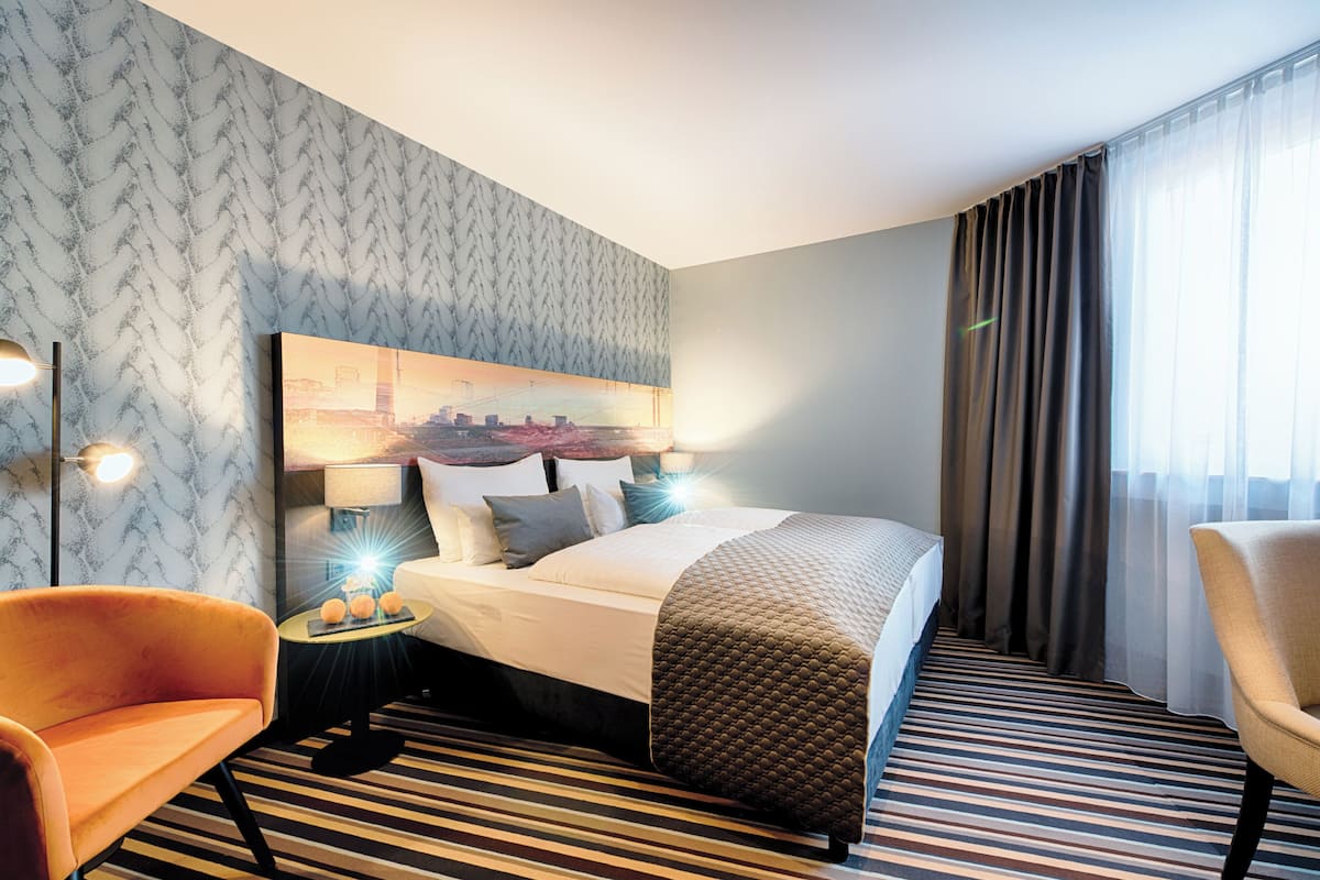 Une chambre d’hôtel avec un lit, deux chaises, une table de chevet avec une lampe, un mur à motifs et une grande fenêtre avec des rideaux.