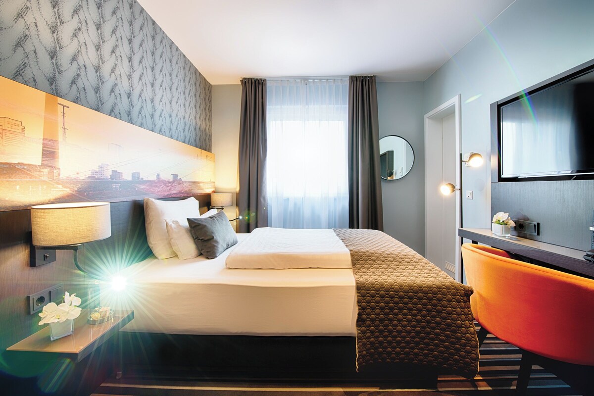 Une chambre d’hôtel avec un lit, une télévision, une chaise et une fenêtre avec des rideaux.