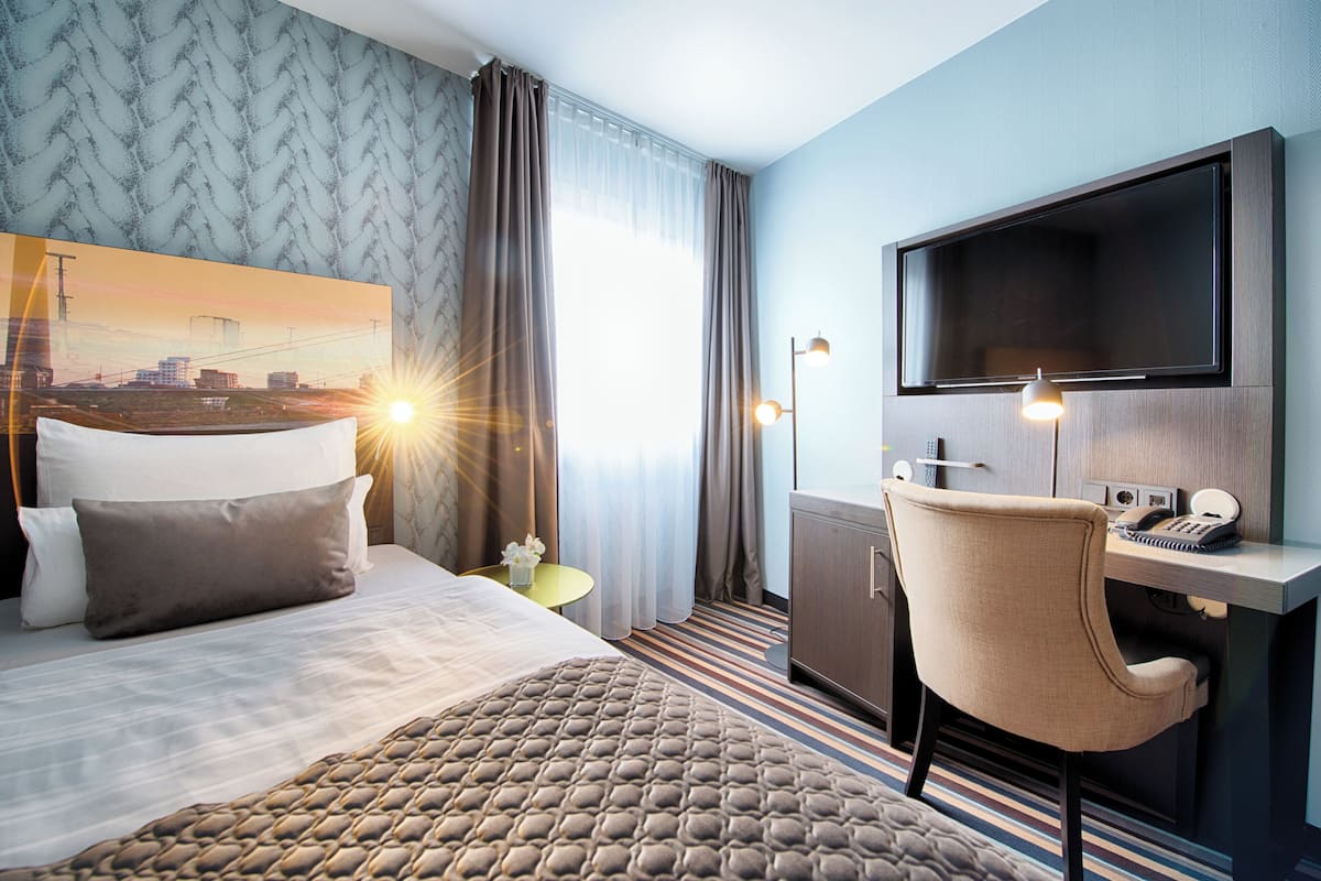 Une chambre d’hôtel équipée d’un lit, d’un bureau, d’une chaise, d’une télévision et d’une grande fenêtre avec des rideaux.