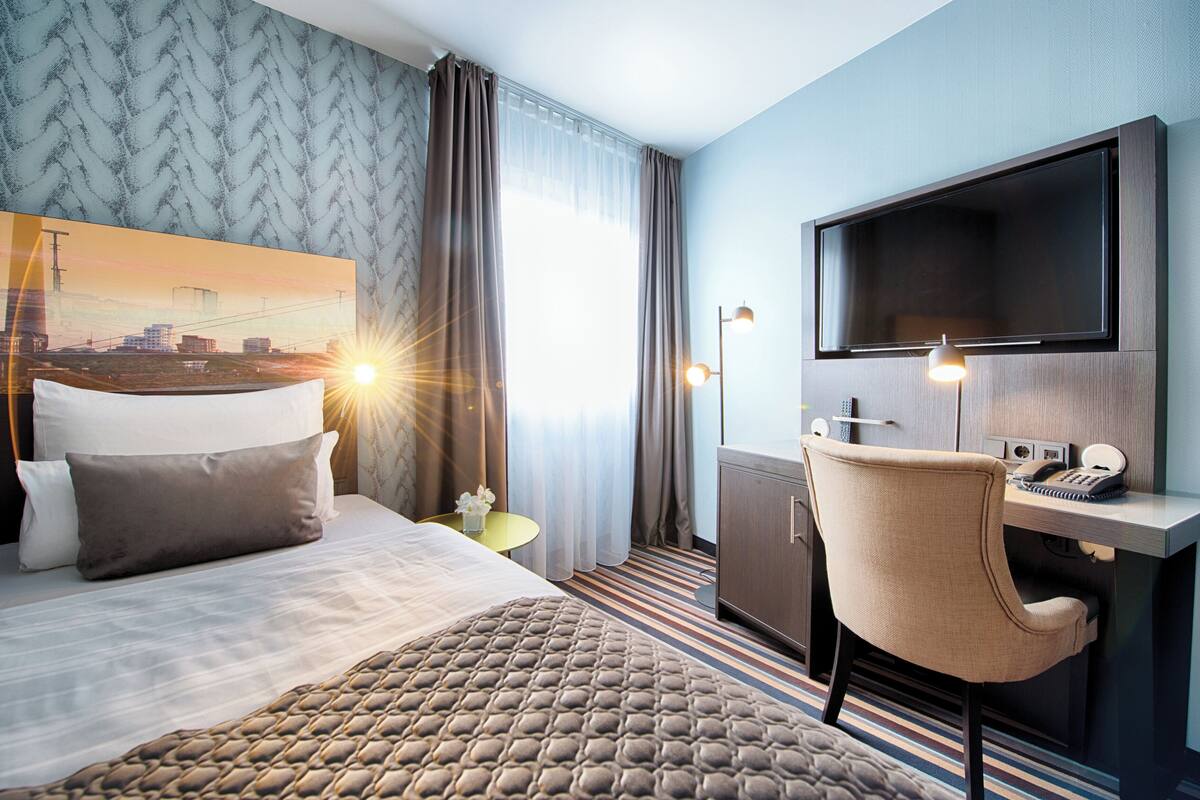 Habitación de hotel con cama, escritorio, silla, televisor y ventana grande con cortinas.