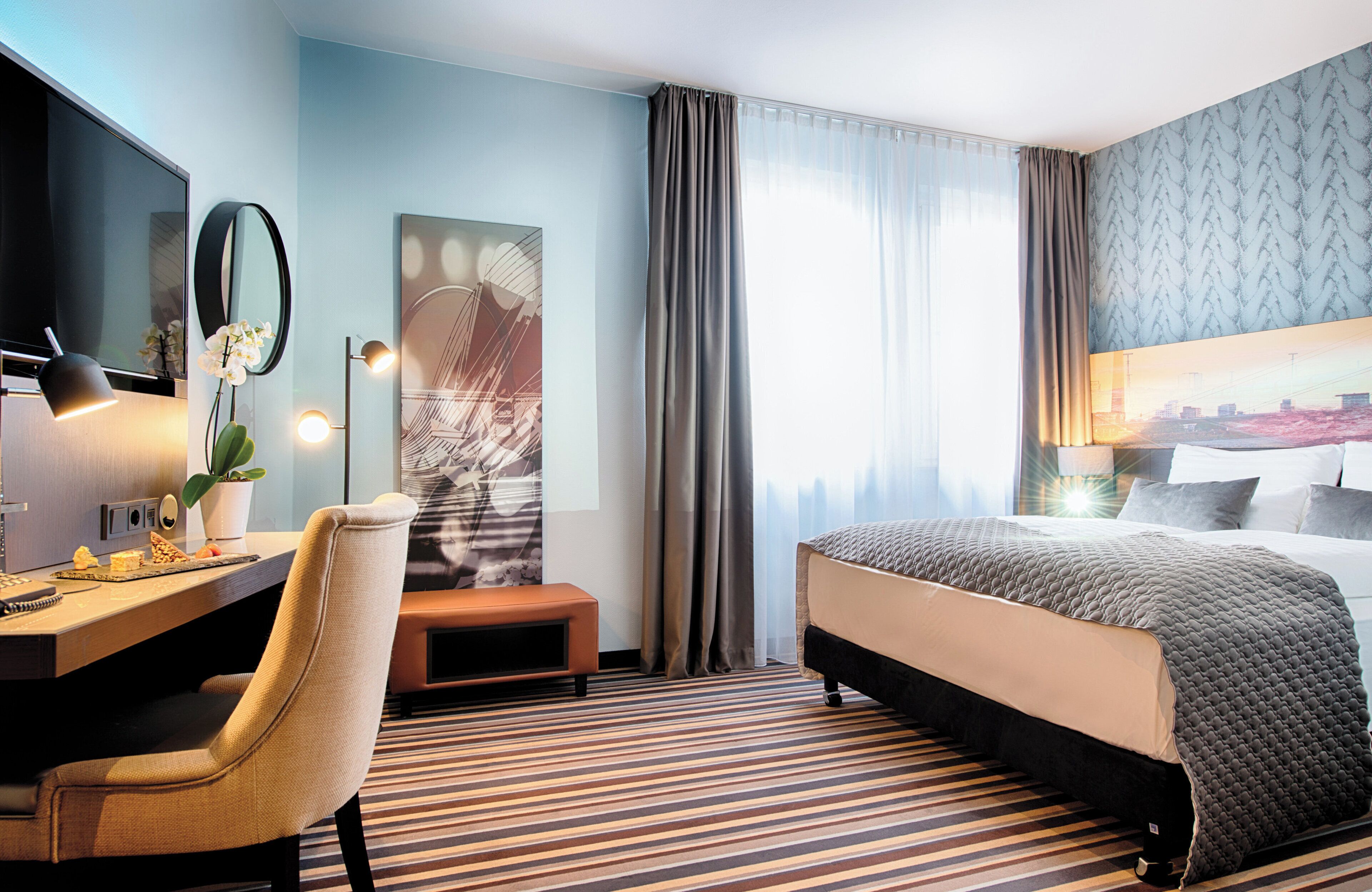 Habitación de hotel con cama, escritorio, silla y una ventana grande con cortinas.