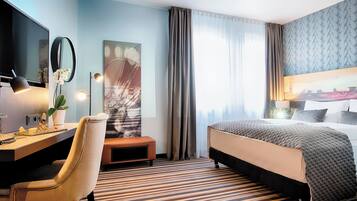 Habitación de hotel con cama, escritorio, silla y una ventana grande con cortinas.