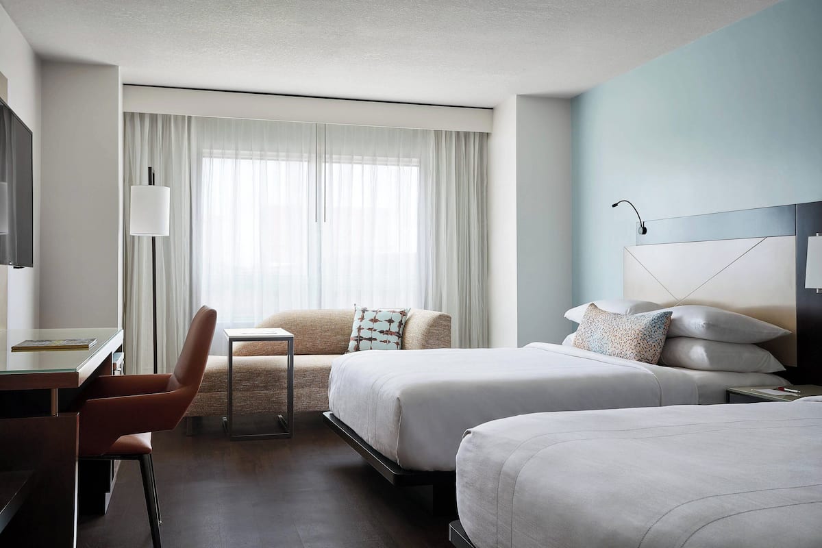 A hotel room with two beds, a desk, a chair, and a large window with curtains.