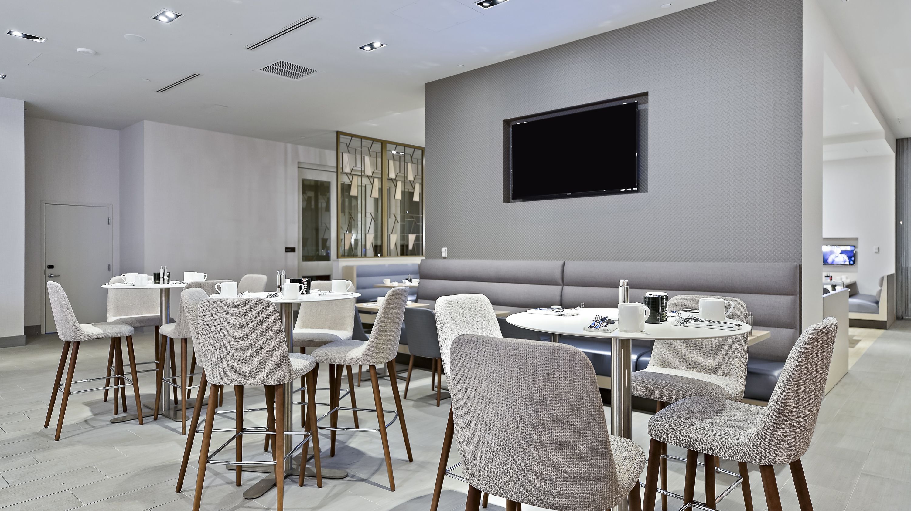 A modern hotel room with a seating area featuring round tables and cushioned chairs, a flat-screen TV mounted on the wall, and a large window providing natural light.