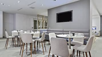 A modern hotel room with a seating area featuring round tables and cushioned chairs, a flat-screen TV mounted on the wall, and a large window providing natural light.