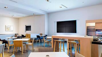 Uma área de refeições moderna com várias mesas e cadeiras, uma cozinha com banquetas de bar, e uma televisão de tela plana grande fixada na parede.