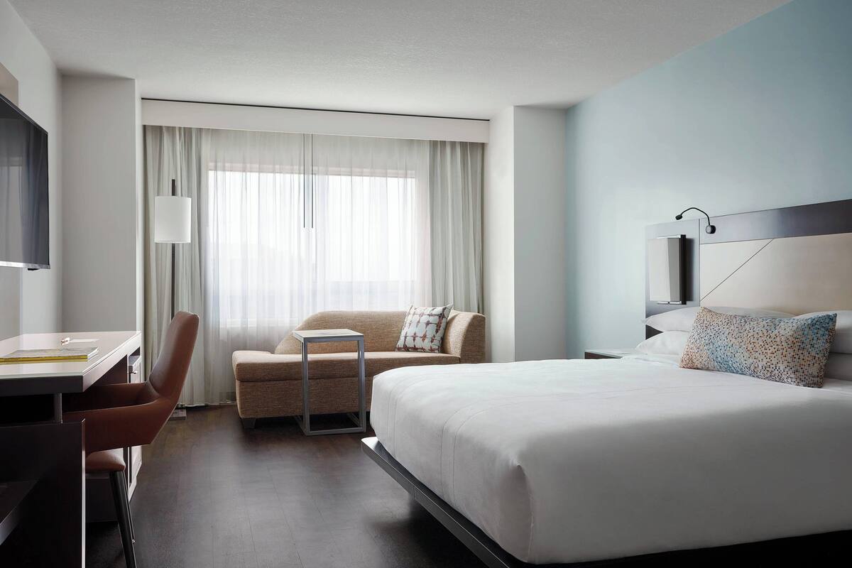 A modern hotel room with a bed, a desk, a chair, a sofa, and a large window with curtains.