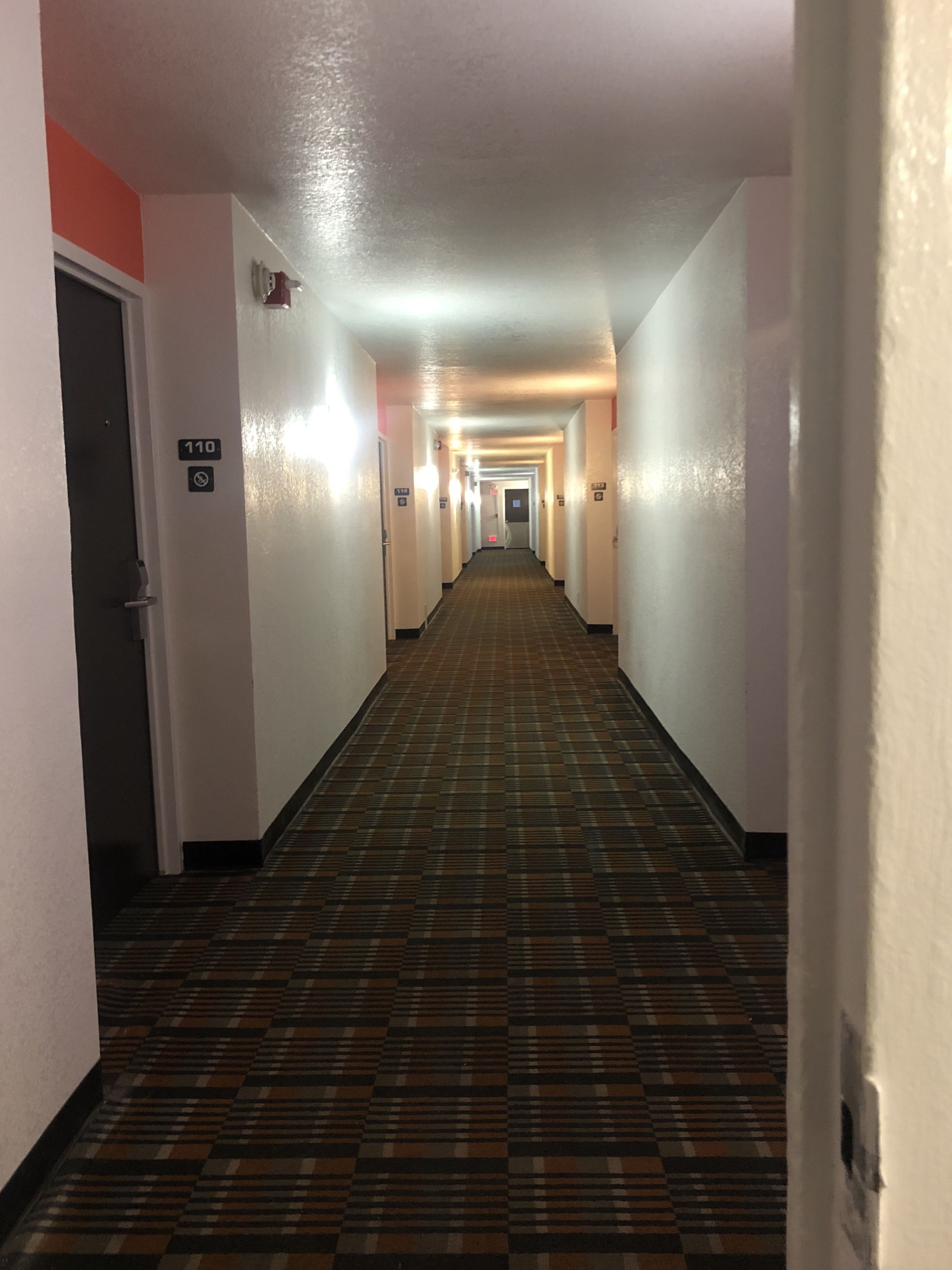 A hotel hallway with a checkered floor, white walls, and doors numbered 110 and 114.