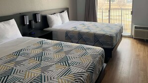 A hotel room with two beds, a nightstand, a wall-mounted alarm clock, and a window with a view of a fenced area. - Motel 6 Lincoln, NE - Airport (Lincoln)