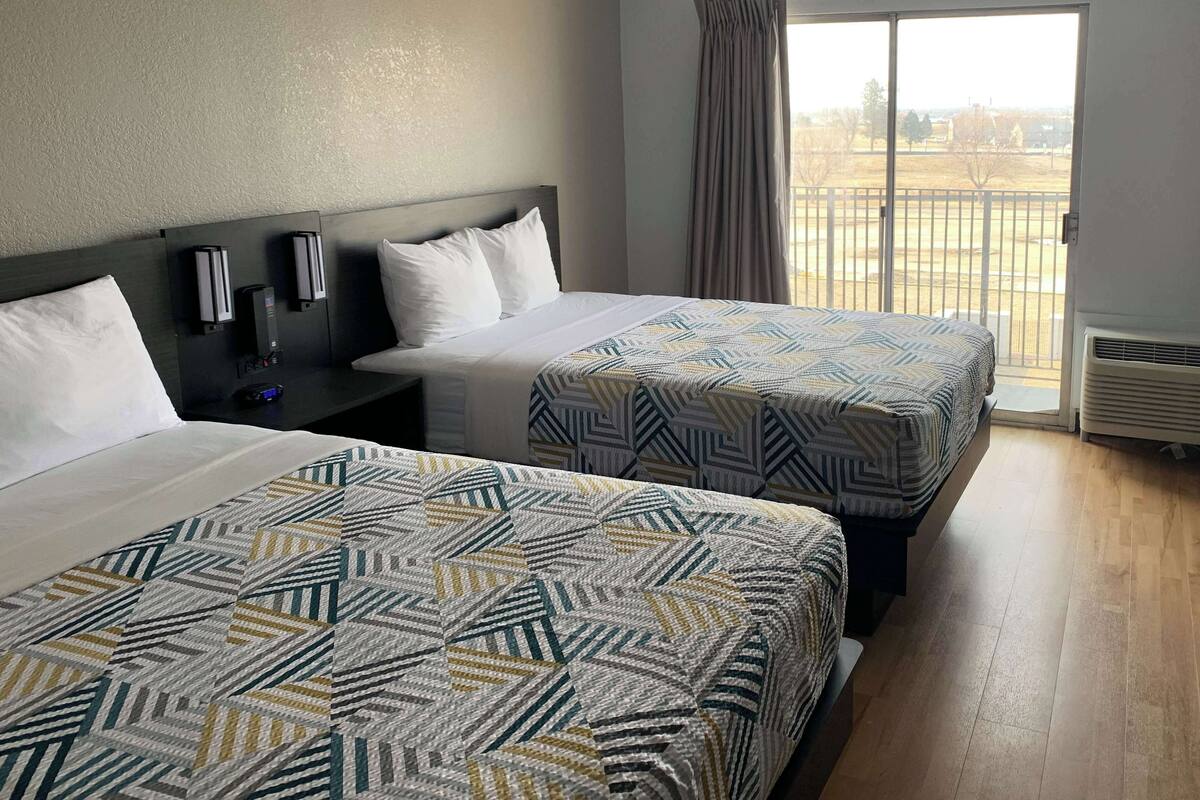 A hotel room with two beds, a nightstand, a wall-mounted alarm clock, and a window with a view of a fenced area.