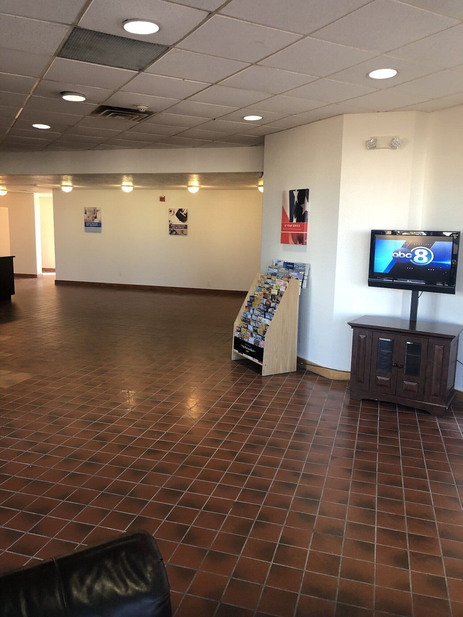 Um hall amplo com piso de azulejos, um balcão de recepção, uma televisão exibindo 'abc 8' e um mural com diversos avisos.