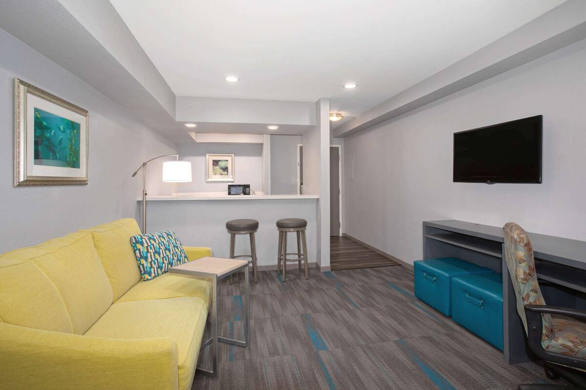 Ein modernes Hotelzimmer mit einem gelben Sofa, einer kleinen Essecke mit Hocker, einem an der Wand befestigten Flachbildfernseher und einem blauen Stauraum.
