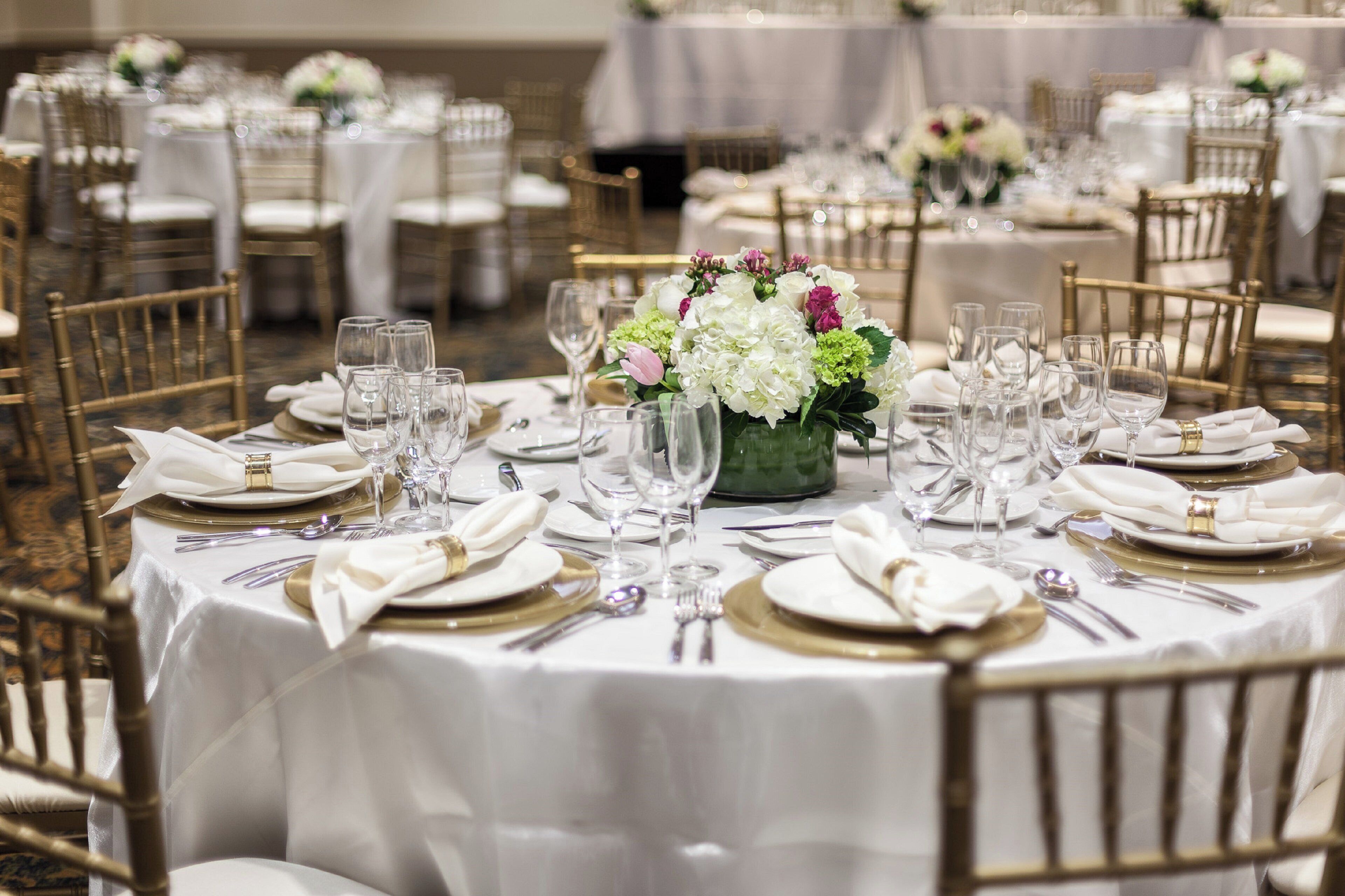 Un salón de banquetes con mesas redondas dispuestas para un evento formal, con manteles blancos, platos con detalles dorados y cristalería transparente.