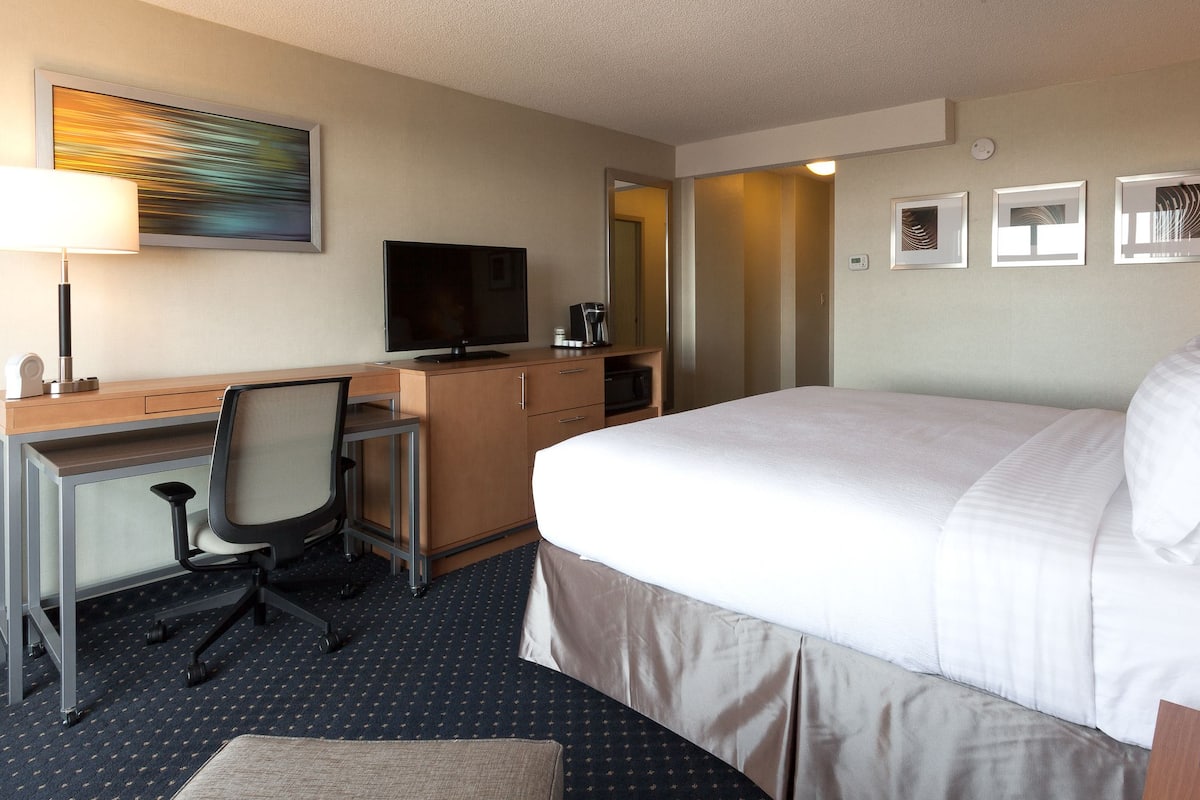A hotel room with a bed, desk, chair, TV, microwave, and artwork on the wall.