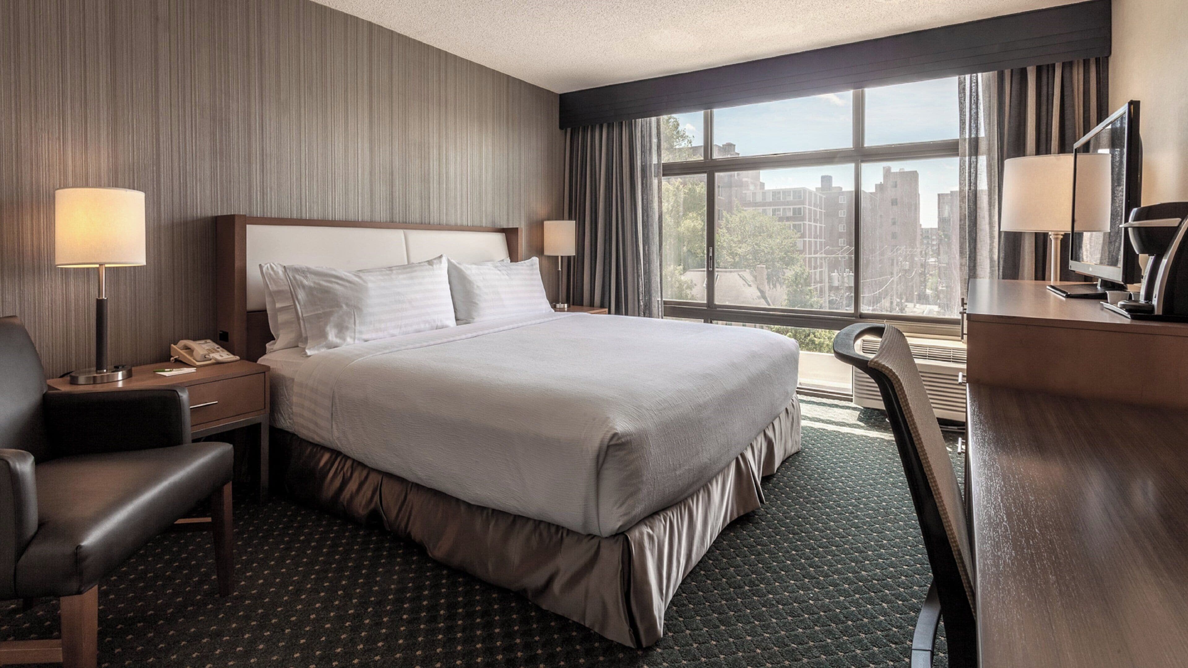 Habitación de hotel con una cama grande, un escritorio, una silla, un teléfono y una ventana con vistas a edificios.