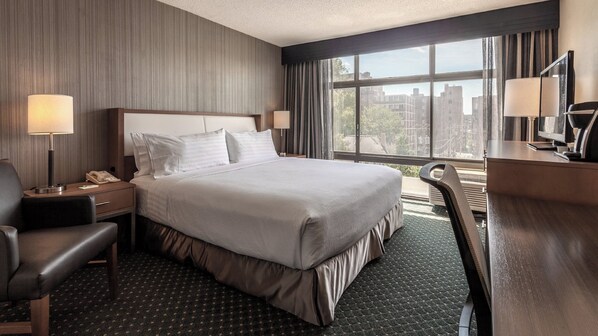 A hotel room with a large bed, a desk, a chair, a telephone, and a window with a view of buildings.
