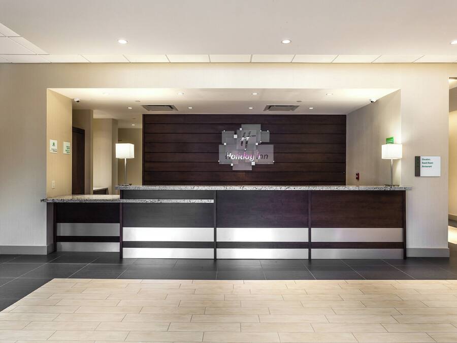 A hotel reception desk with a modern design, a digital sign displaying 'Holiday Inn', and a sign indicating the location of elevators, board room, and restaurant.
