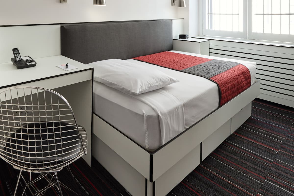 A modern hotel room with a bed, desk, chair, and a window with blinds.