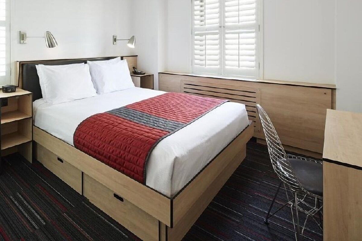 Une chambre d’hôtel comprenant un lit, des tables de chevet, un bureau, une chaise et une fenêtre avec des stores.