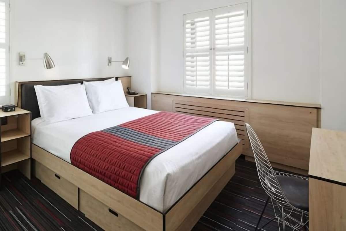 A hotel room with a bed, bedside tables, a chair, a desk, and a window with blinds.
