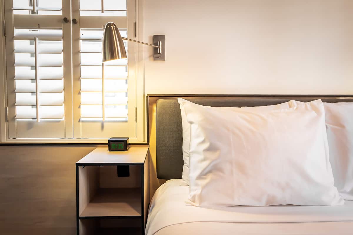 A neatly made bed with white linens and a single white pillow, a bedside table with a lamp, and a window with blinds.