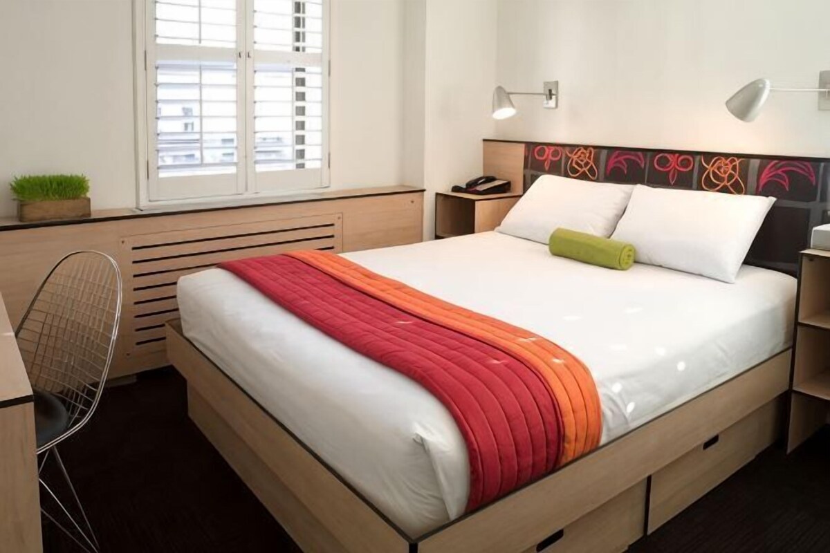 Une chambre d’hôtel avec un lit, une table de chevet, une lampe et une fenêtre avec des stores.