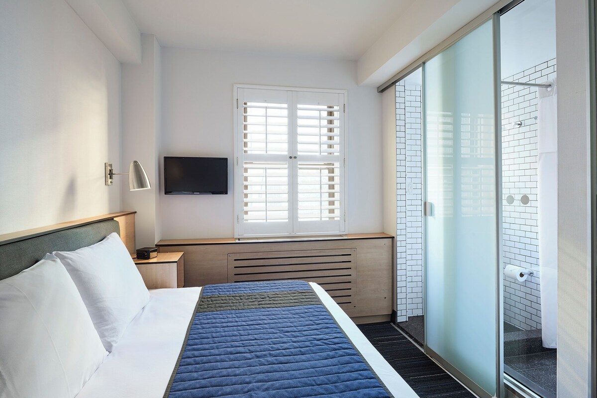 Une chambre d’hôtel avec un lit, une télévision, une fenêtre avec des stores et une vue sur l’extérieur.