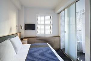 Habitación de hotel con cama, televisor, ventana con persianas, radiador y puerta corrediza de vidrio a un balcón.