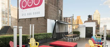 A rooftop patio with colorful Adirondack chairs, red cushioned seating, and a sign for 'The Pod Hotel' with a contact number.