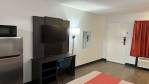A compact hotel room with a kitchenette, a flat-screen TV mounted on the wall, a desk with a chair, and a door with a red curtain. - Motel 6 Macclenny, FL (MacClenny)