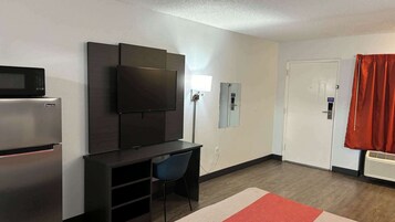 Habitación de hotel compacta con kitchenette, televisor de pantalla plana montado en la pared, escritorio con silla y puerta con cortina roja.