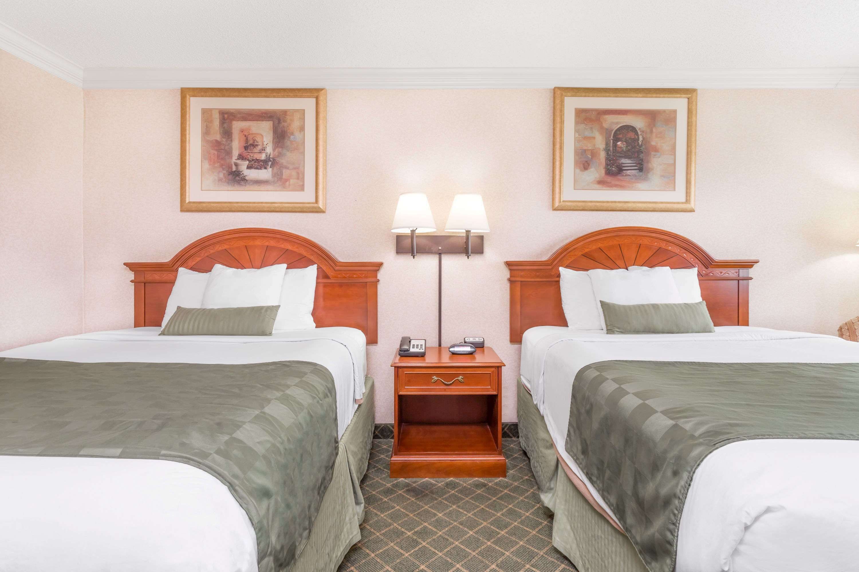 A hotel room with two beds, each with a wooden headboard, a nightstand with a lamp, and a small drawer. There are two framed pictures on the wall above the beds.
