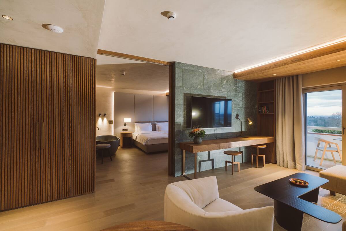 A modern hotel room with a large bed, a desk with two chairs, a sofa, and a TV mounted on a stone wall.