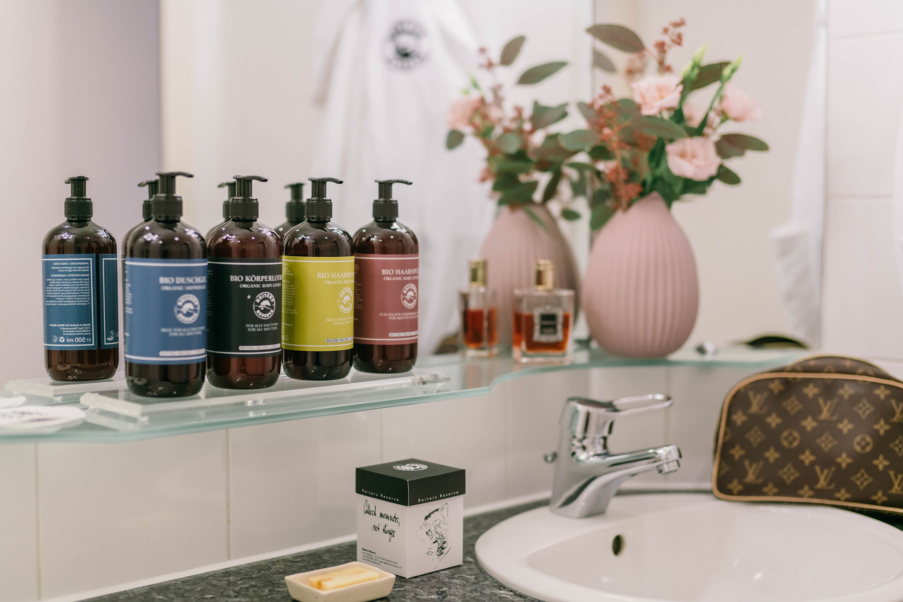 Ein Badezimmer mit einer Glasablage, auf der verschiedene Flaschen mit Haarpflegeprodukten stehen, einem weißen Waschbecken und einer Vase mit rosa Blumen.