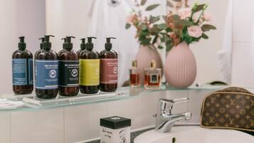Ein Badezimmer mit einer Glasablage, auf der verschiedene Flaschen mit Haarpflegeprodukten stehen, einem weißen Waschbecken und einer Vase mit rosa Blumen.
