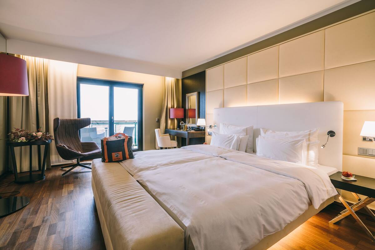 A hotel room with a large bed, a desk, a chair, and a view of the city through the window.