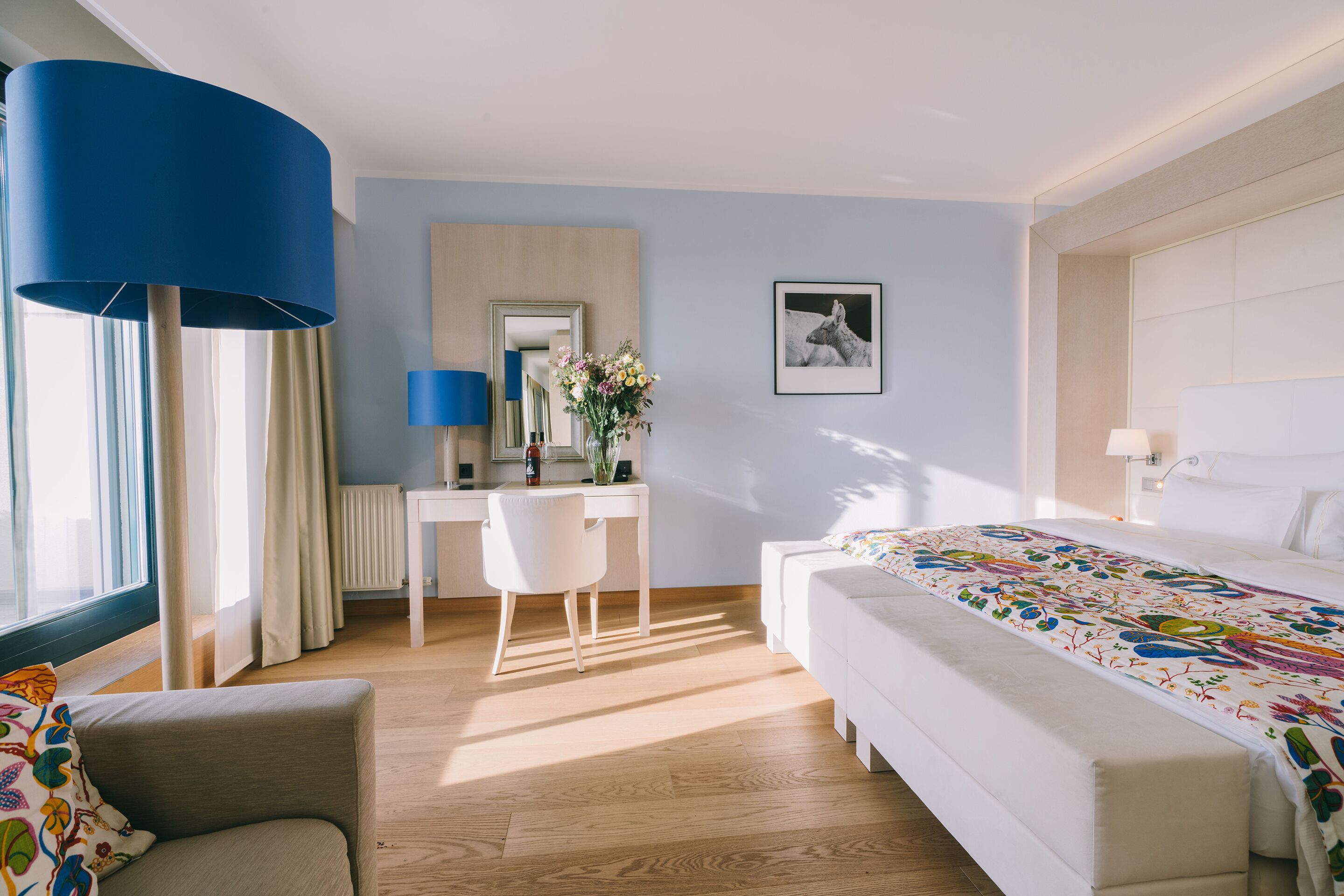 Ein Hotelzimmer mit Bett, Schreibtisch, Stuhl, großem Fenster mit Vorhängen und einer Wandlampe.