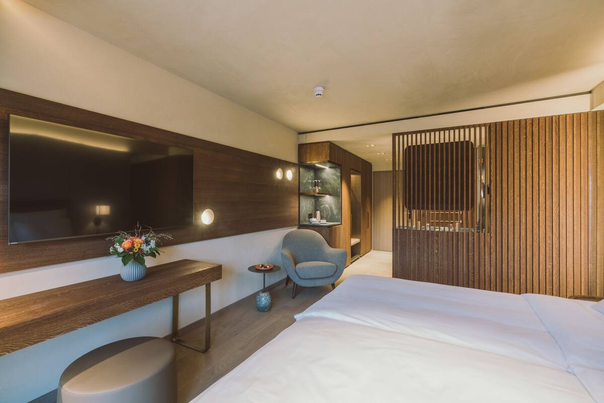 A modern hotel room with a large bed, a seating area with a chair and a small table, a vanity with a mirror, and a wooden partition.