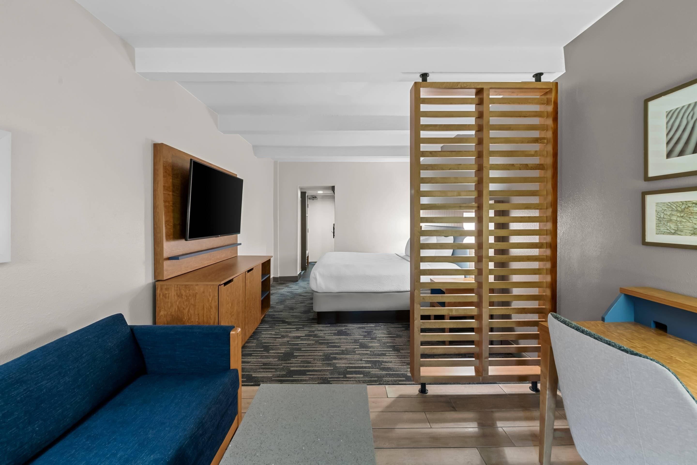 A hotel room with a blue sofa, a TV on a wooden stand, a bed, a desk, and a wooden partition.