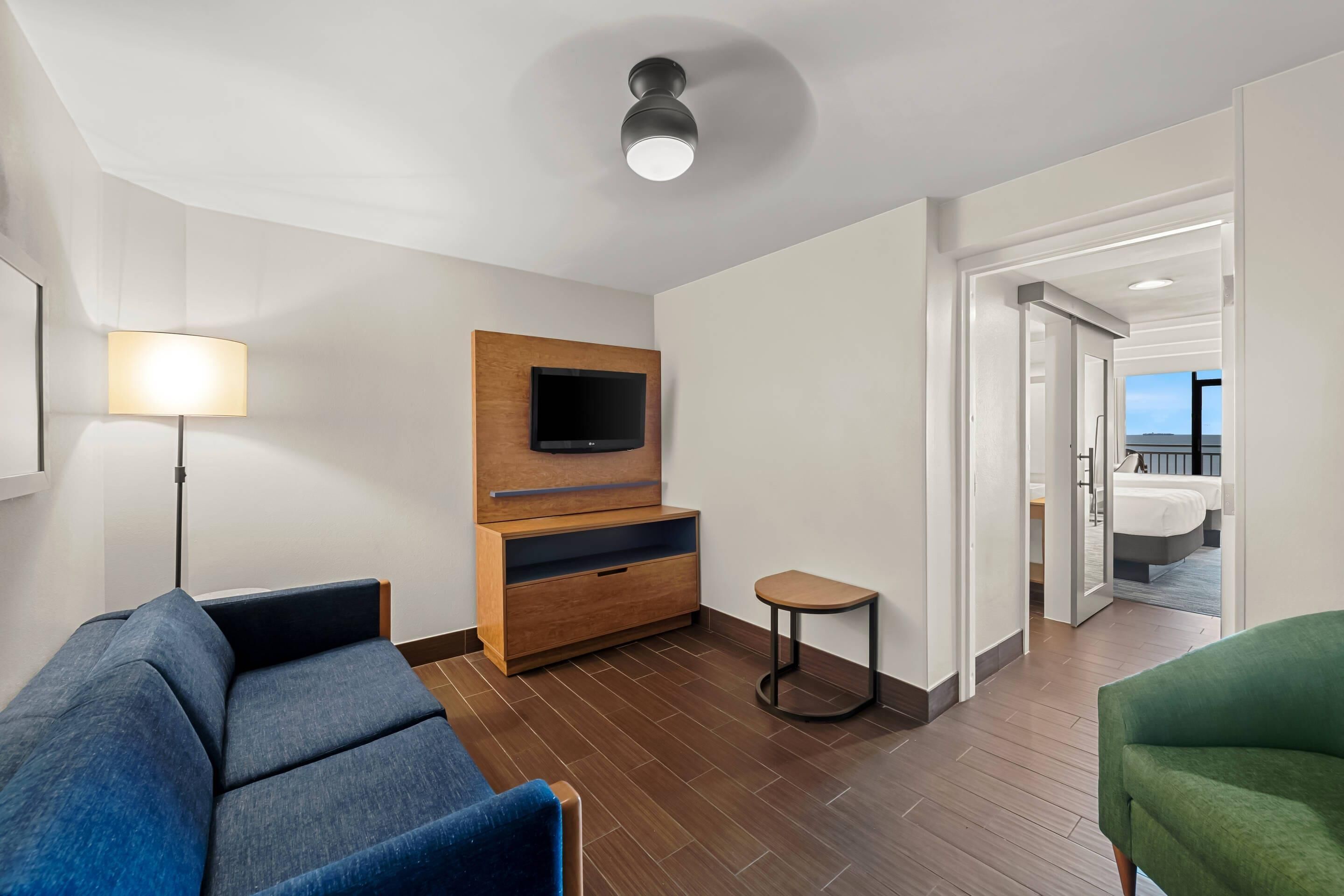 Una camera d'albergo moderna con un mobiletto in legno, una televisione a schermo piatto, un divano blu, una poltrona verde e un tavolino rotondo.