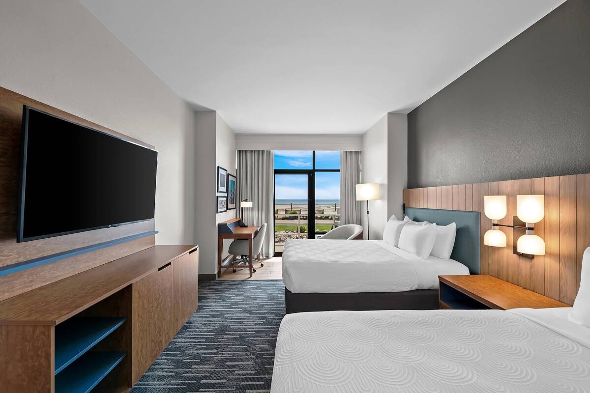 Une chambre d’hôtel avec deux lits, un bureau, une télévision et vue sur la plage.