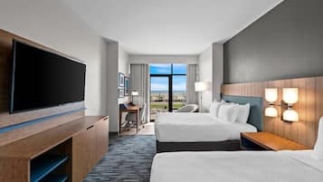 A hotel room with two beds, a desk, a TV, and a view of the beach.
