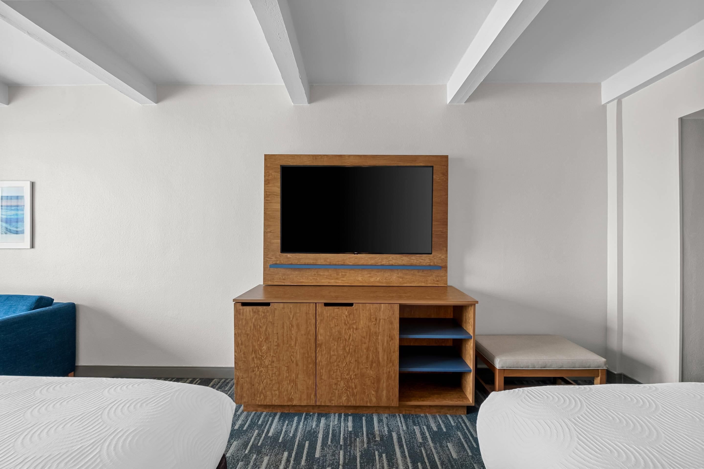 A hotel room with a large TV on a wooden stand, two beds, a small bench, and a framed picture on the wall.