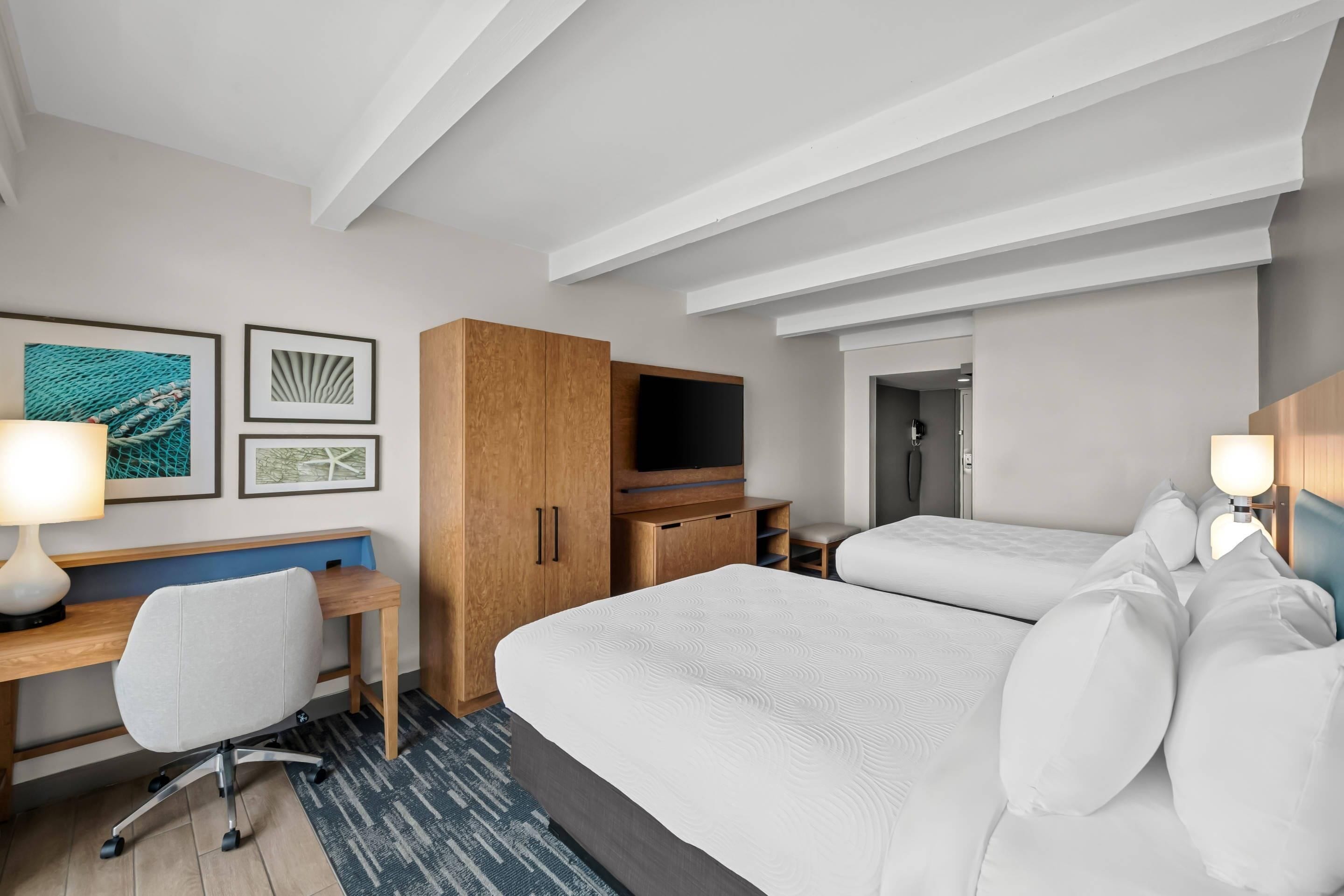 A hotel room with a bed, a desk, a chair, a TV, and artwork on the wall.