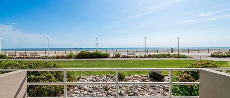 A patio area with a view of the beach, featuring a railing, seating, and a pathway leading to the beach.