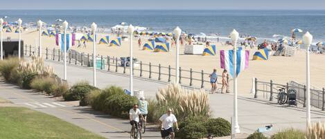 A coastal promenade with a beach in the background, featuring a paved walkway, lamp posts, and beach umbrellas.