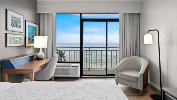 A hotel room with a bed, a desk, a chair, a lamp, and a view of the ocean through a large window.