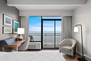 A hotel room with a bed, a desk, a chair, a lamp, and a view of the ocean through a large window. - Country Inn & Suites by Radisson, Virginia Beach (Oceanfront) (Virginia Beach)