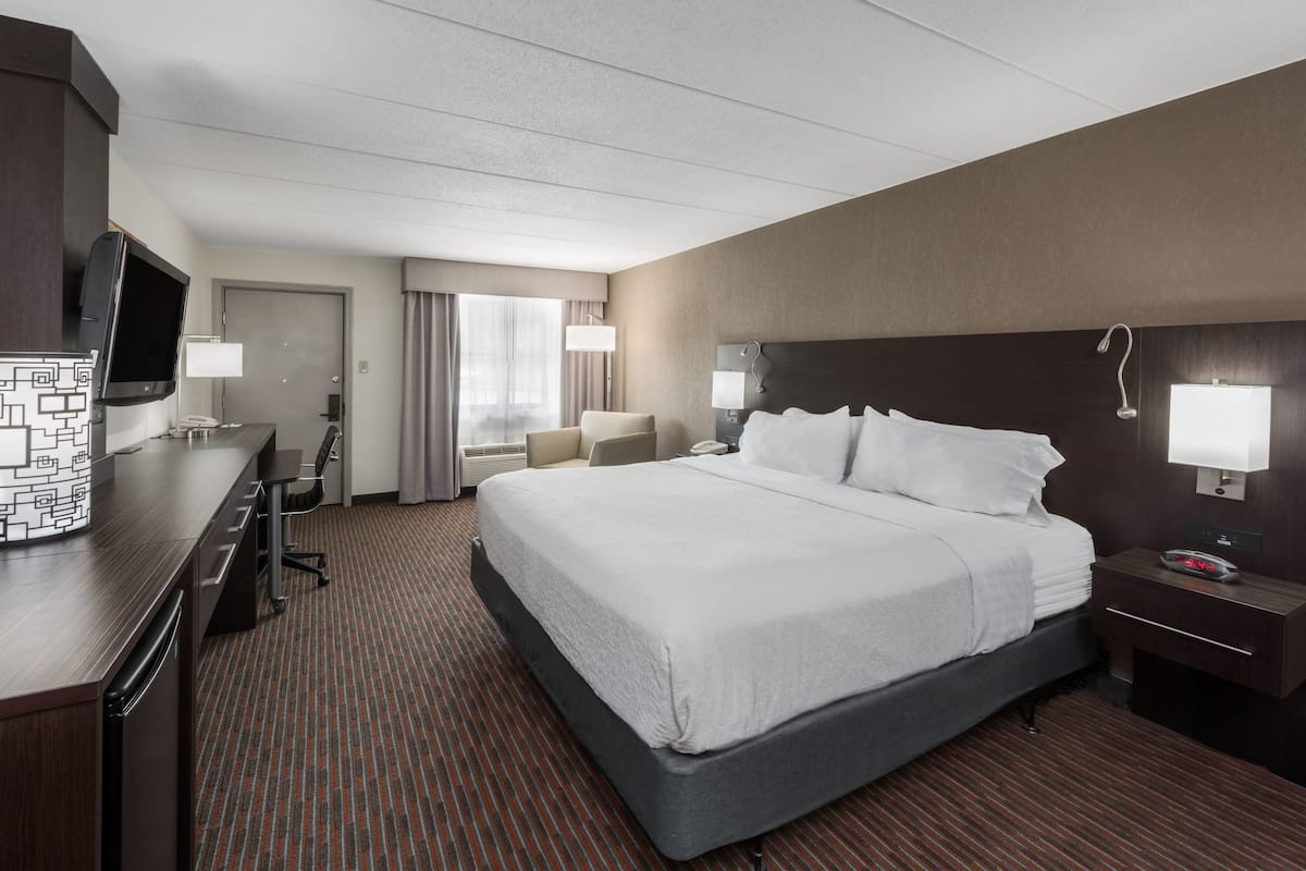 A hotel room with a large bed, a desk with a flat-screen TV, a chair, a desk lamp, and a nightstand with a clock.
