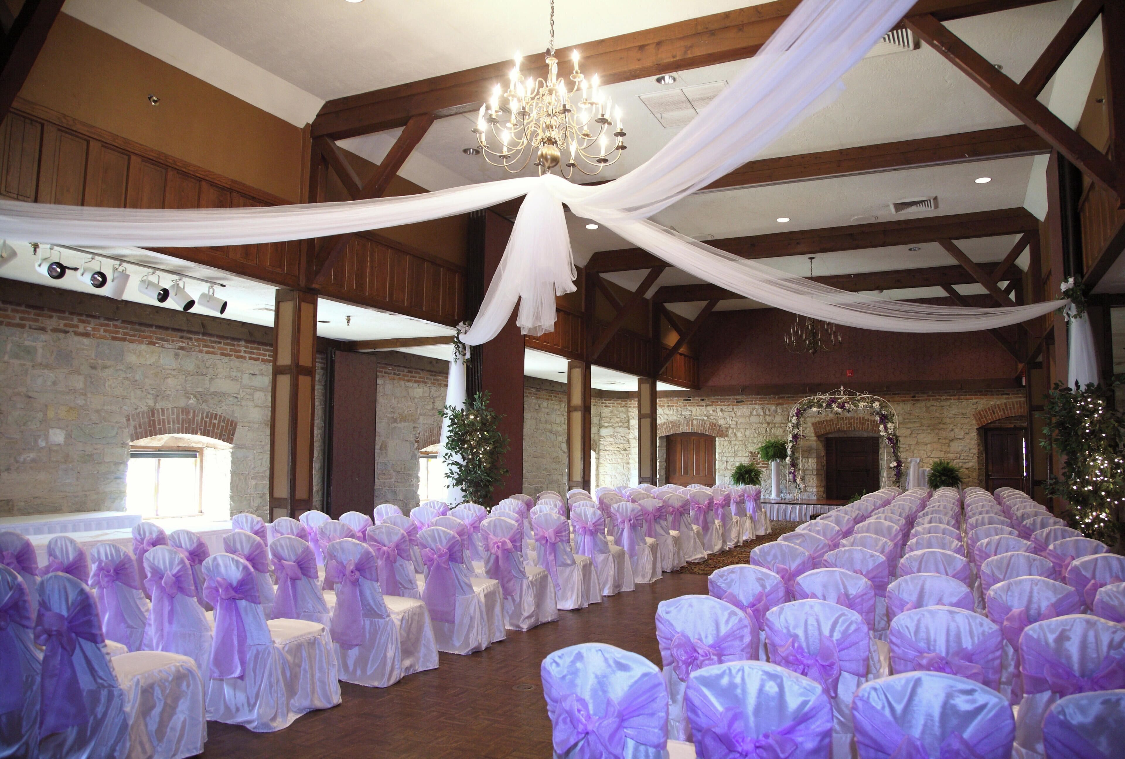 Un salón de banquetes con filas de sillas cubiertas de tela blanca y morada, un candelabro y una pared de piedra.